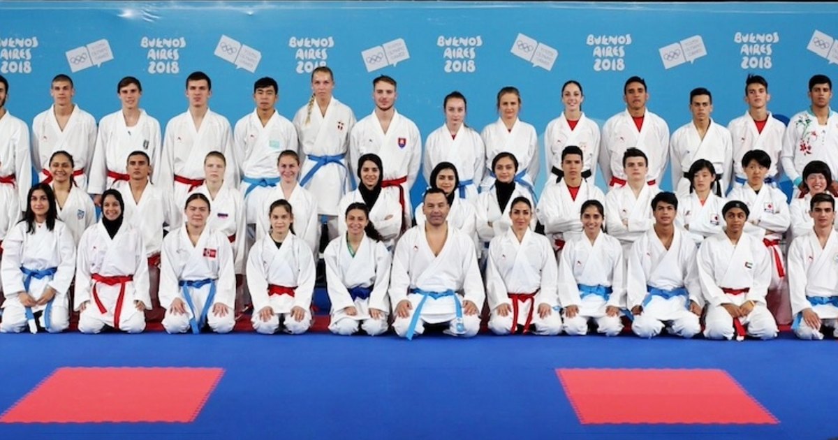 Jeunes karatékas en karategi blanc, ceintures rouges et bleues, agenouillés et debout sur un tatami bleu et rouge devant un fond aux couleurs de Buenos Aires 2018